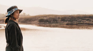 Woman wearing a hat standing by calm water, looking toward the horizon.